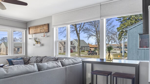 Un soggiorno moderno con un divano modulare grigio, una ventola a soffitto e un'ampia finestra che si affaccia su un quartiere residenziale.