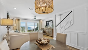 Un salon moderne avec une table ronde en bois, des canapés de couleur beige, un lustre en osier et une vue sur l’océan grâce à de grandes fenêtres.