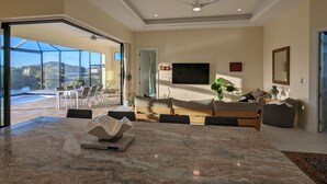 A spacious living room with a large marble table, a ceiling fan, a flat-screen TV, and a view of a pool and outdoor area. - New home on Gulf access water near beaches! (Cape Coral)