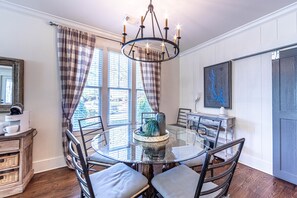 Uma sala de jantar com mesa de vidro rodeada por cadeiras, um lustre e uma janela com cortinas.