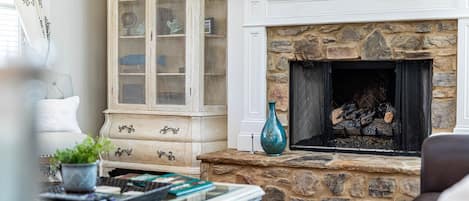 Una sala de estar con chimenea de piedra, un armario blanco con puertas de cristal y un jarrón azul sobre la repisa.