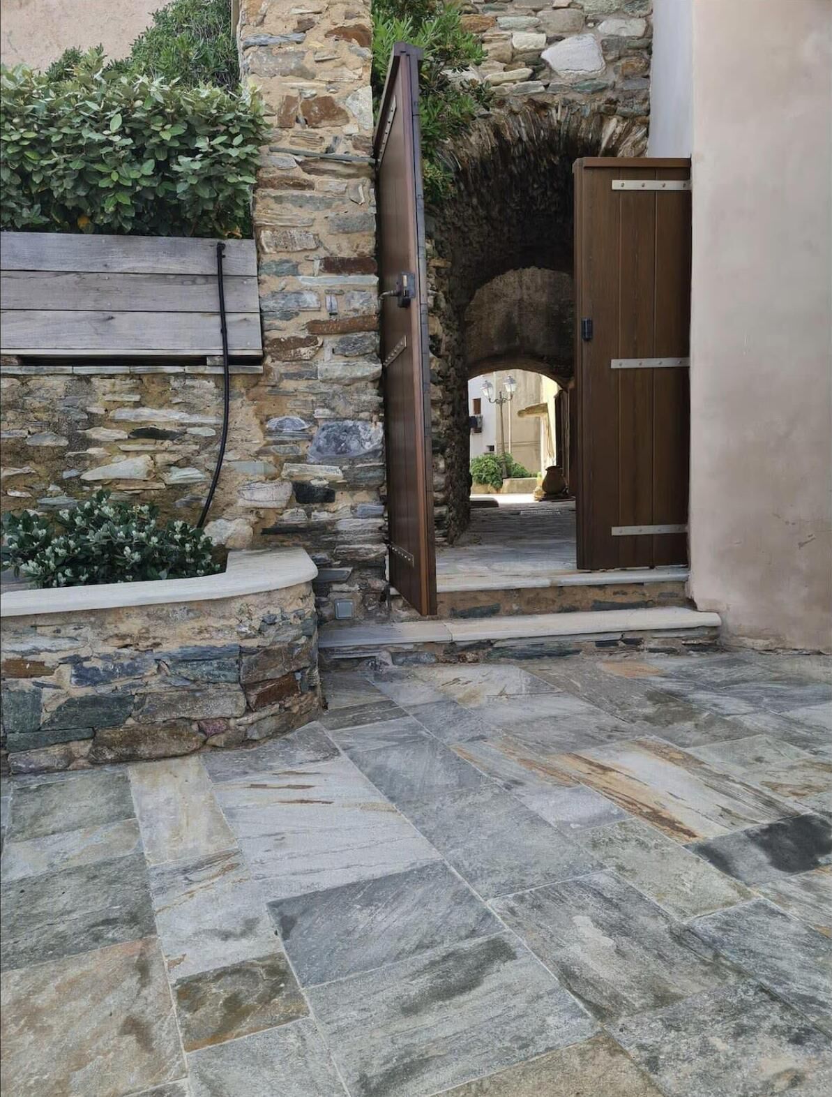 A stone courtyard with a wooden door leading to an archway, surrounded by stone walls and a wooden bench.