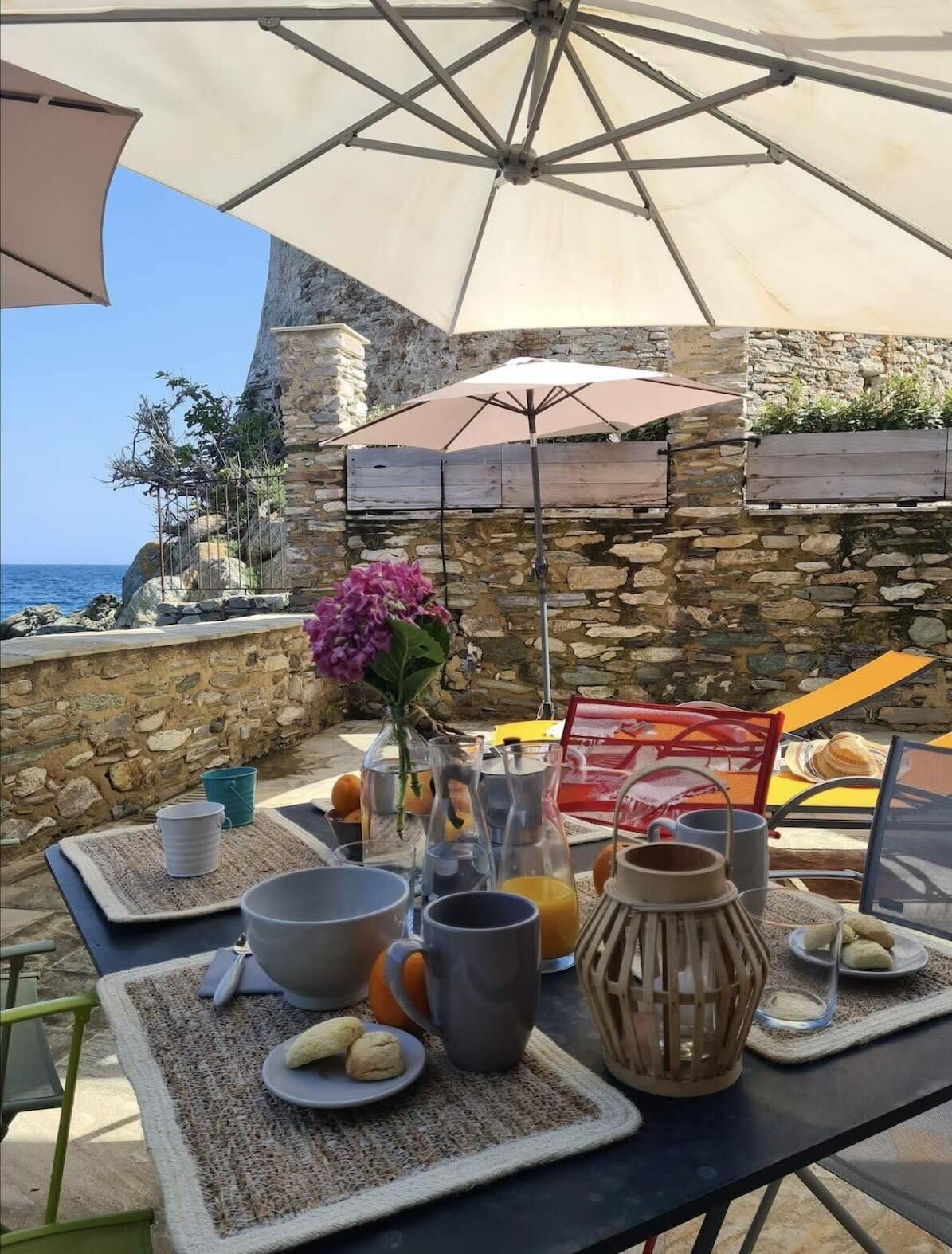 A breakfast setup with a table, drinks, and food items on a terrace with a stone wall and ocean view.