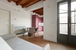 Un soggiorno moderno con un divano, un tavolo da pranzo con sedie, un angolo cottura con mobili rosa e un balcone con ringhiera metallica.