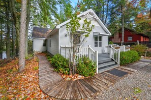 A white cabin with a wooden deck and a small porch, surrounded by trees and a body of water.