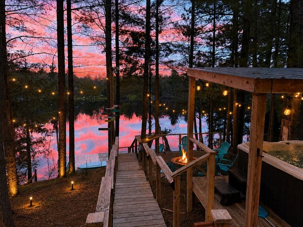 A wooden deck with a fire pit, outdoor seating, and a hot tub overlooking a lake at dusk.