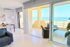 A modern living room with a sofa, dining table, and a glass door leading to a balcony with a view of the sea.