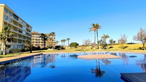 A swimming pool with a clear blue sky, a multi-story hotel building, and palm trees.