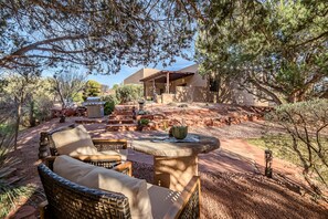 Relax in a serene outdoor space with a barbecue grill and wicker seating area surrounded by lush plants and trees.