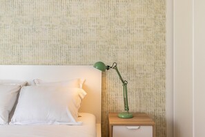 Una cama bien hecha con ropa de cama y almohadas blancas, una lámpara de escritorio verde sobre una mesita de noche de madera, y un papel tapiz con textura en la pared.