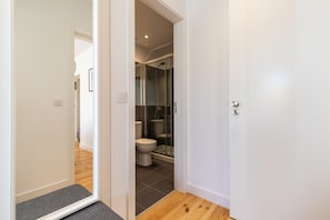 Un couloir étroit mène à une salle de bain équipée de toilettes, d’une douche et d’un espace de douche séparé par des parois en verre.