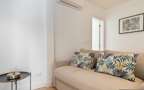 Un salon minimaliste avec un canapé beige, deux coussins à motifs, une petite table ronde agrémentée d’un objet décoratif et un climatiseur fixé au mur.