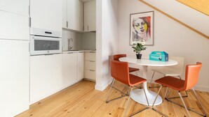 Una cocina moderna con armarios blancos, horno empotrado y comedor con mesa blanca y sillas naranjas.