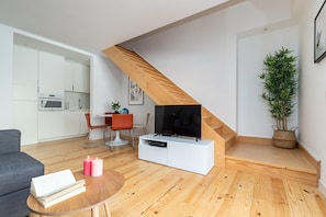 Una sala de estar moderna con una escalera de madera, un televisor de pantalla plana sobre un mueble blanco, un comedor con mesa y sillas, y una cocina al fondo.