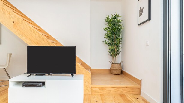 Una sala moderna con una televisión de pantalla plana sobre un mueble blanco, una escalera de madera, una planta en maceta y un cuadro en la pared.