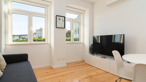 Ett modernt vardagsrum med stora fönster, en platt-TV, ett vitt matbord och en grå soffa.