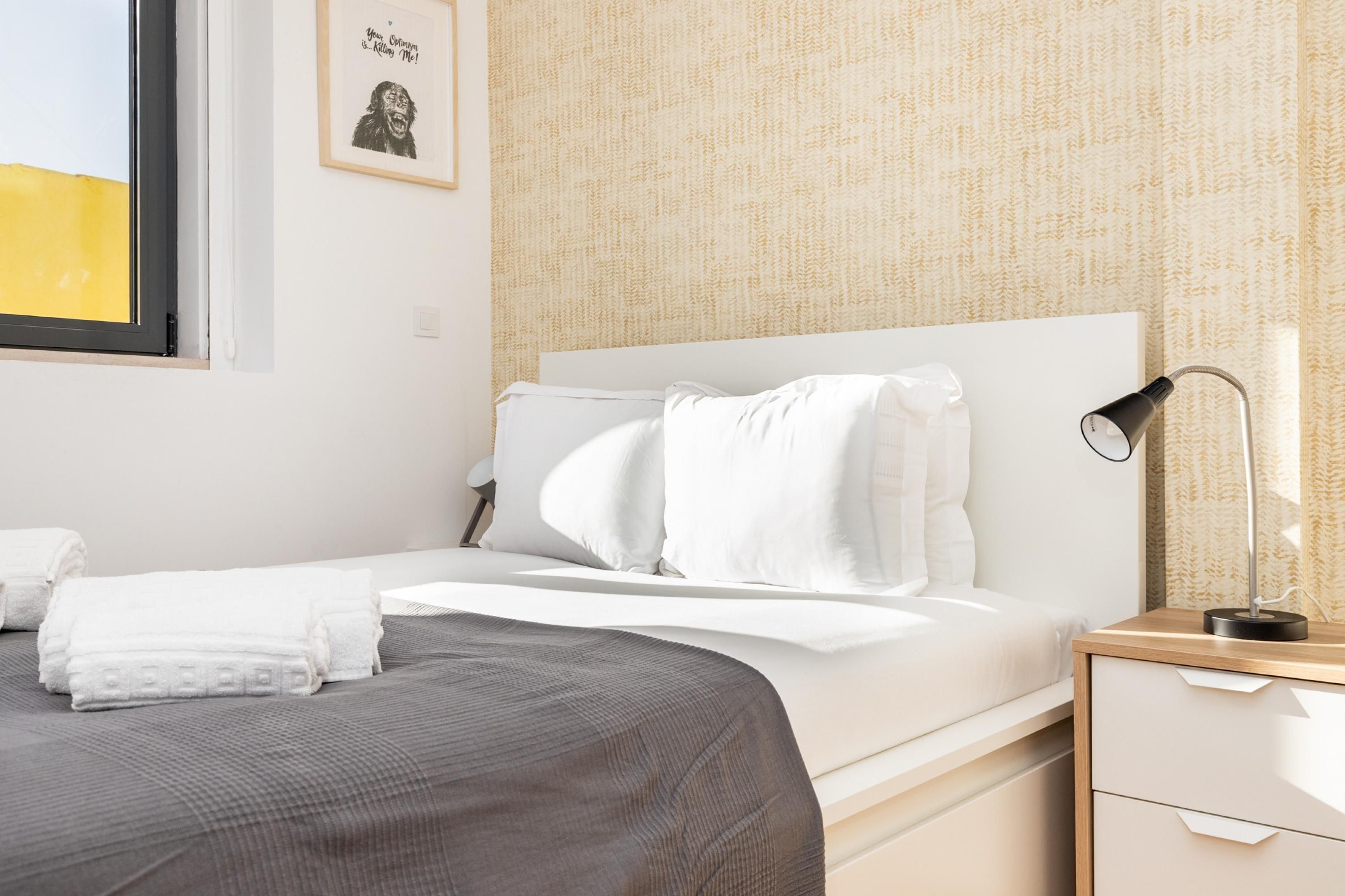 Cama bem arrumada com lençóis brancos, luminária de cabeceira e quadro na parede.
