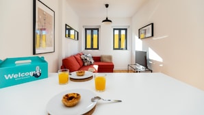 Ein modernes Wohnzimmer mit einem weißen Tisch, einem roten Sofa und einer blauen 'Welcome'-Box. An der Wand hängen zwei gerahmte Bilder und eine Deckenleuchte.