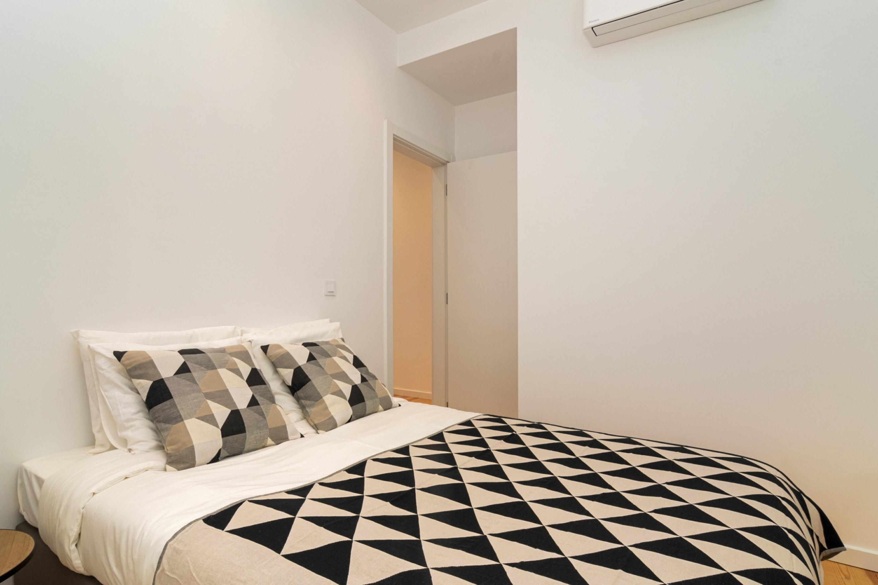 A bedroom with a bed, two pillows, a bedside table, and a door leading to another room.