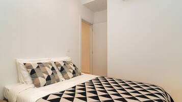 A bedroom with a bed, two pillows, a bedside table, and a door leading to another room.