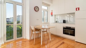 A modern kitchen with white cabinets, a wooden dining table, and chairs. There is a clock on the wall and a fire alarm sign. - Arroios Concept III by Homing (Lisbon)
