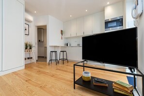 Ein modernes Wohnzimmer mit einem großen Flachbildfernseher, einem Couchtisch mit Büchern und einer Vase, sowie einer Küchenzeile mit weißen Schränken und einer Mikrowelle.