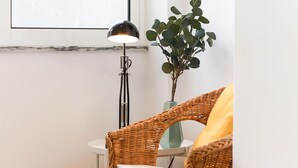 A wicker chair with a white cushion, a floor lamp, and a potted plant in a corner. - Lisbon Typical by Homing (Lisbon)