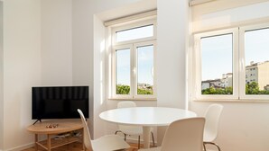Una sala de estar moderna con una mesa redonda blanca, sillas blancas, un televisor de pantalla plana y ventanas amplias que ofrecen vistas de la ciudad.