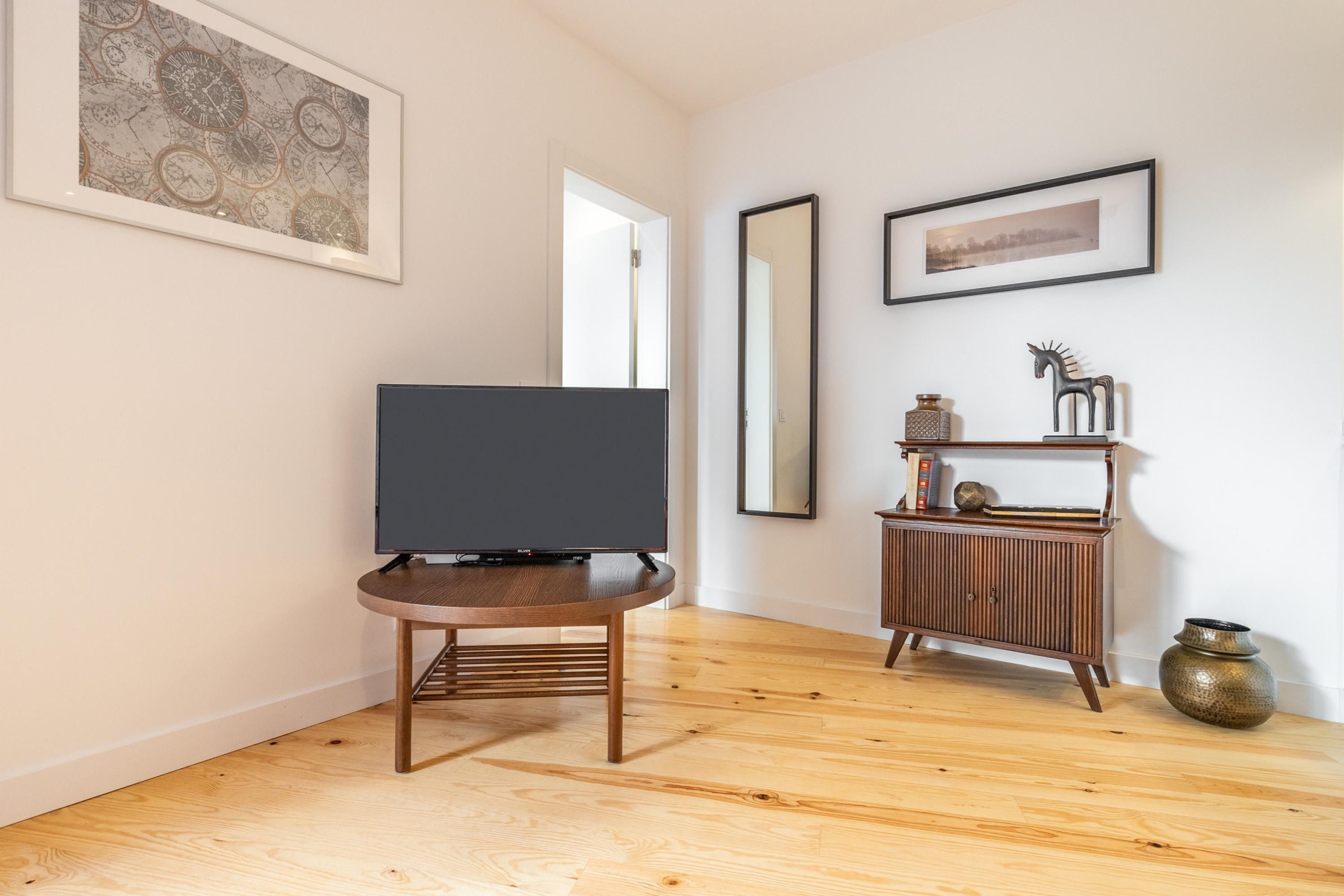 A modern living room with a flat-screen TV on a wooden stand, a wooden cabinet with decorative items, and a large framed picture on the wall.