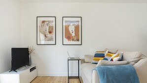 A modern living room with a flat-screen TV on a white entertainment unit, a glass coffee table, a beige sofa with a blue throw blanket, and two framed abstract paintings on the wall. - Belem Design 13 by Homing (Lisbon)