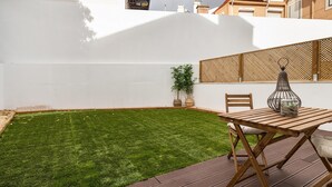 Eine Terrasse mit einem Holztisch und Stühlen, ein kleiner Gartenbereich und eine weiße Wand.