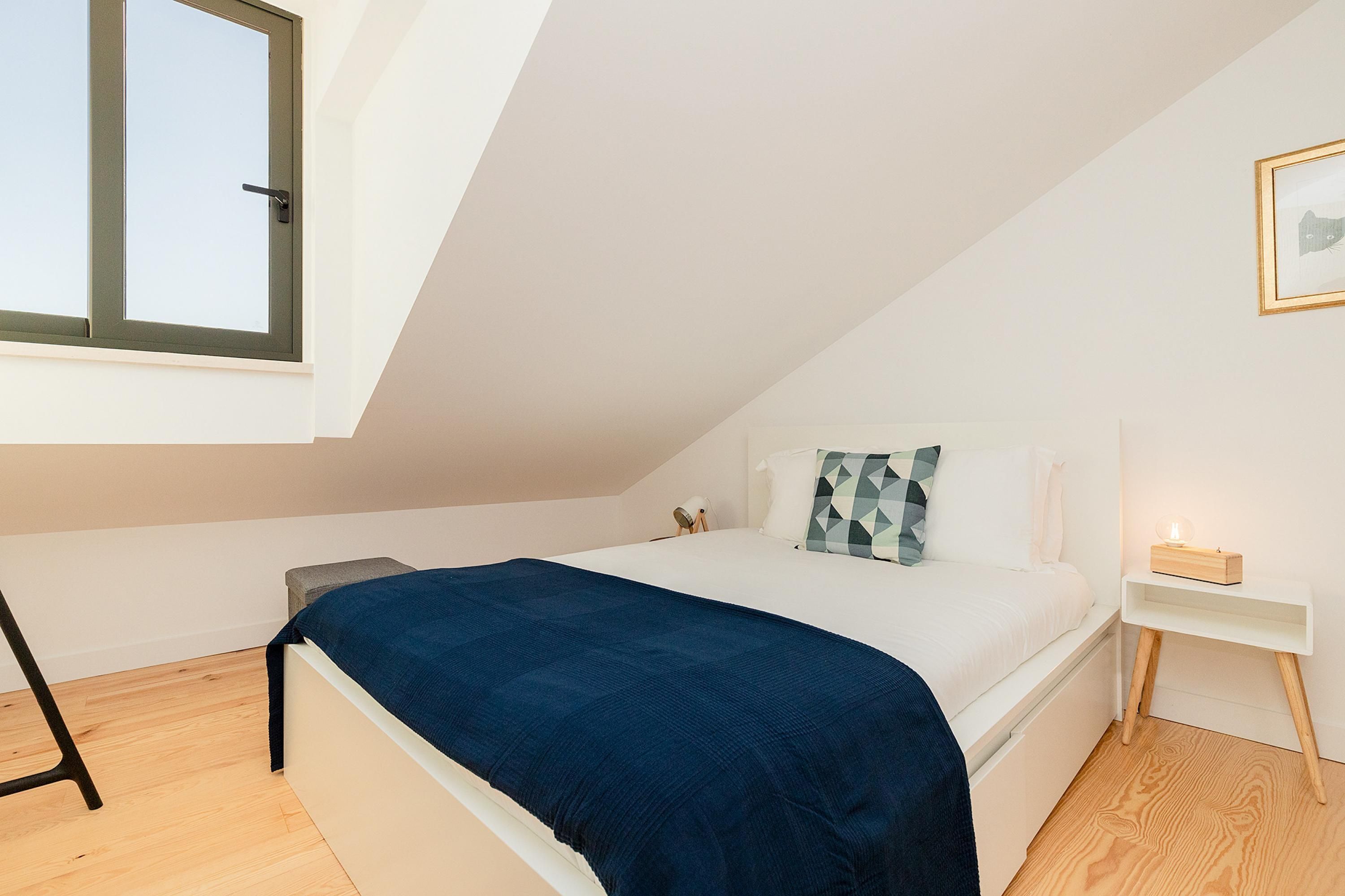 A bedroom with a bed, a bedside table, a nightstand, a window, and a picture on the wall.
