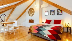 Une chambre avec un plafond en pente, un lit recouvert d’une courtepointe colorée, un bureau avec une chaise et une petite table avec une lampe.