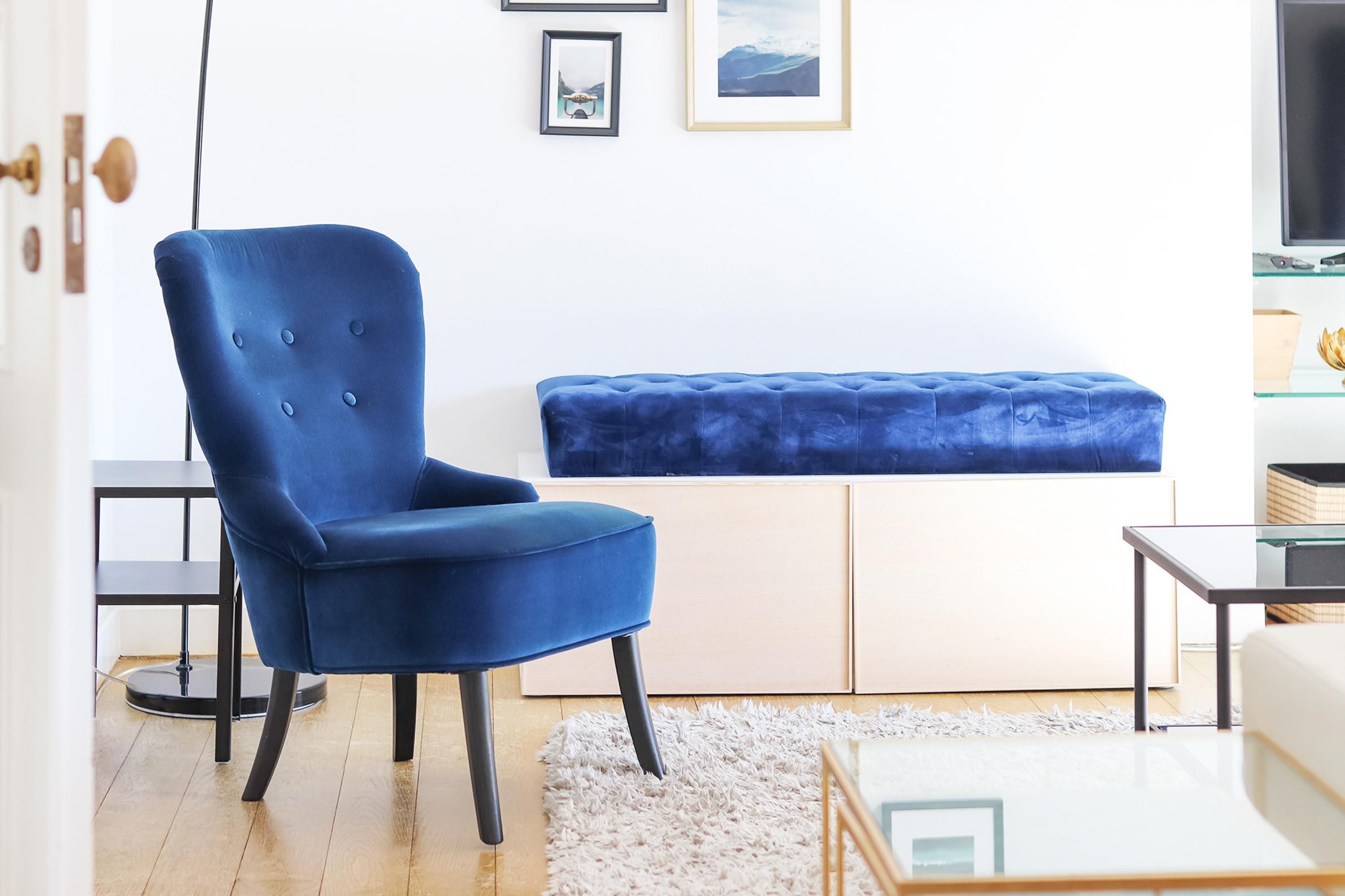 A modern living room with a blue armchair, a tufted bench, a glass coffee table, and a flat-screen TV on a stand.