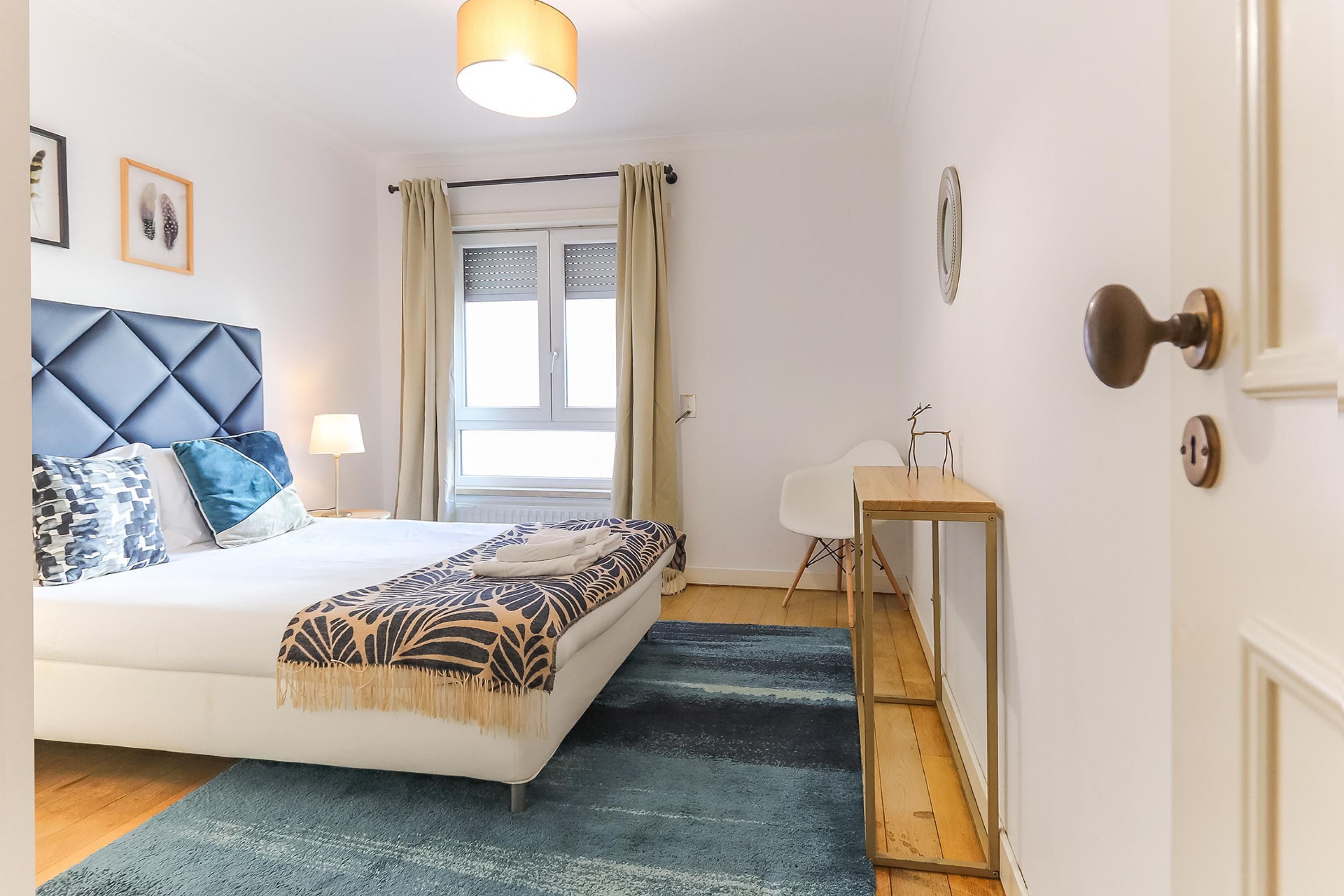Une chambre moderne avec un lit, une table de chevet, une lampe, une fenêtre avec des rideaux et une porte avec une poignée.