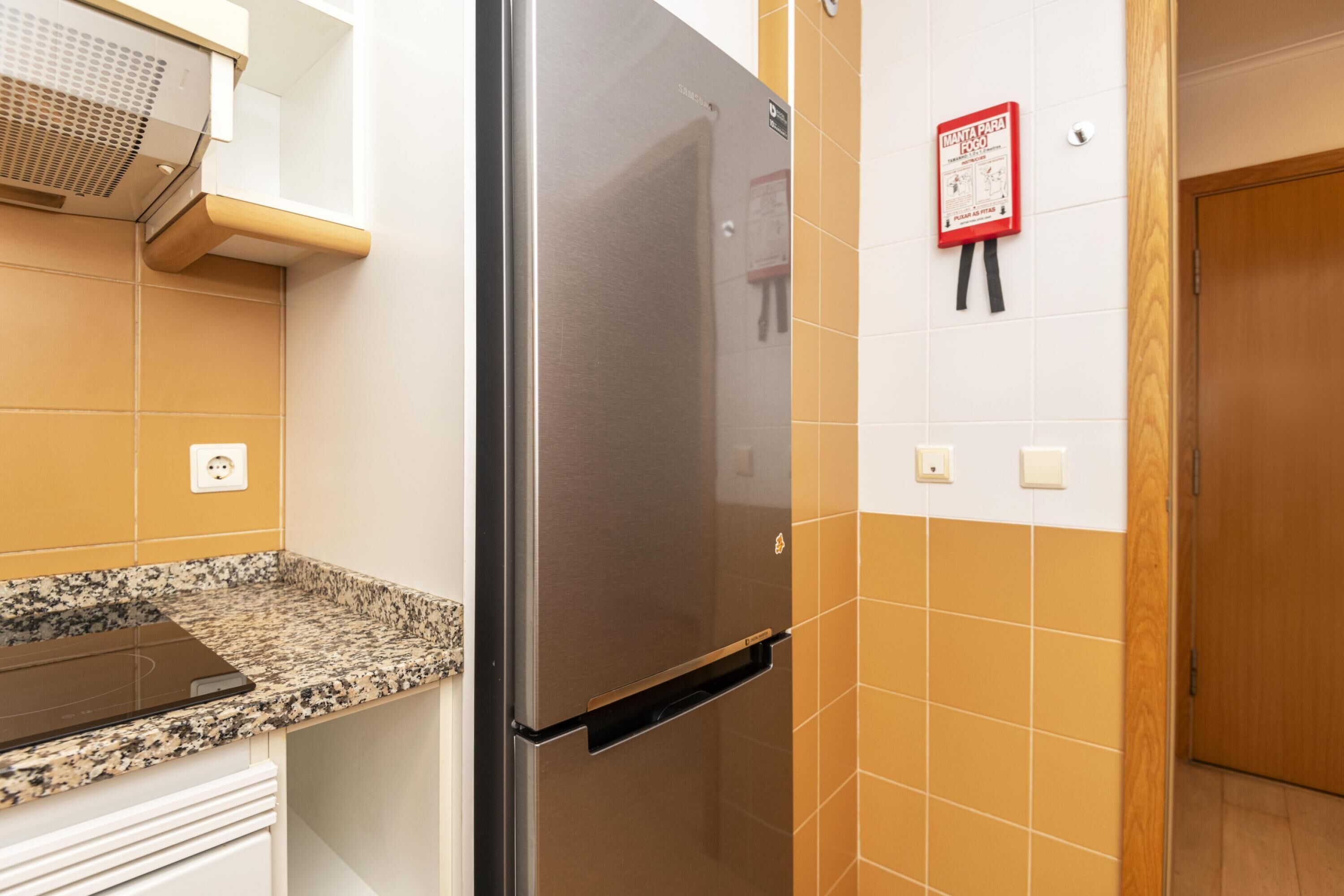 A kitchen with a stainless steel refrigerator, a granite countertop, and a wall-mounted fire alarm.
