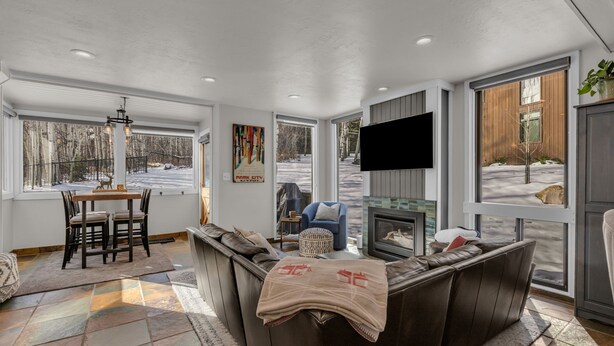 A modern living room with a stone tile floor, a brown leather sofa, a blue armchair, a fireplace, and large windows overlooking a snowy landscape. - Cozy Townhome next to PC Resort! Stylish 2BR/2BA +loft, deck, +more (Park City)