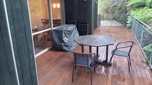An outdoor patio with a covered table and chairs, a wooden deck, and a glass door leading to an interior space with chairs and a table. - Holiday house on 1 acre of native bush a few minutes walk to lake Brunner (Moana)