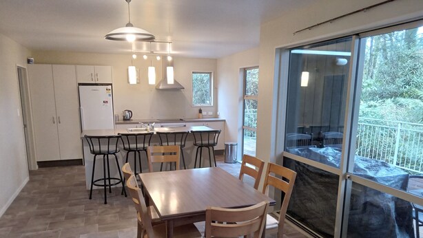 A modern kitchen with a dining area, featuring a large table and chairs, a refrigerator, and a window with a view of greenery. - Holiday house on 1 acre of native bush a few minutes walk to lake Brunner (Moana)