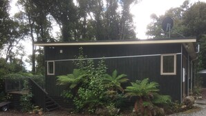 A small, dark-colored building with a gravel driveway, surrounded by lush greenery and a metal staircase leading up to a deck. - Holiday house on 1 acre of native bush a few minutes walk to lake Brunner (Moana)