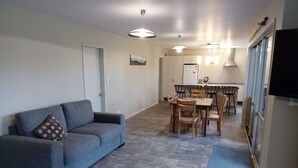 A living room with a sofa and a dining table with chairs. There is a kitchen area with a refrigerator and a sink. - Holiday house on 1 acre of native bush a few minutes walk to lake Brunner (Moana)