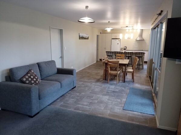 A living room with a sofa and a dining table with chairs. There is a kitchen area with a refrigerator and a sink.
