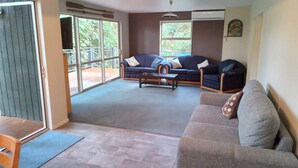 A spacious living room with a large window, a sofa, a coffee table, and a dining area. - Holiday house on 1 acre of native bush a few minutes walk to lake Brunner (Moana)