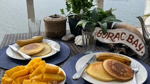 Ein Tisch ist reichhaltig mit Frühstück zubereitet, darunter Pfannkuchen, Mangostücke und ein Glas Wasser, mit Blick auf einen See und Hügel im Hintergrund.