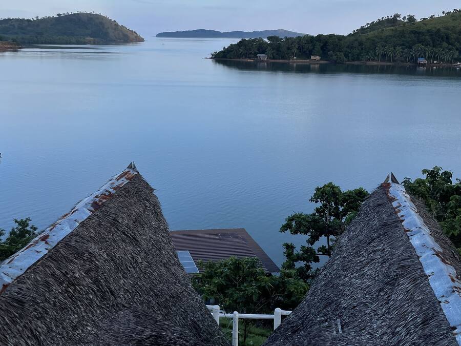 Vista de un lago tranquilo con una estructura de techo de paja en primer plano y colinas distantes.