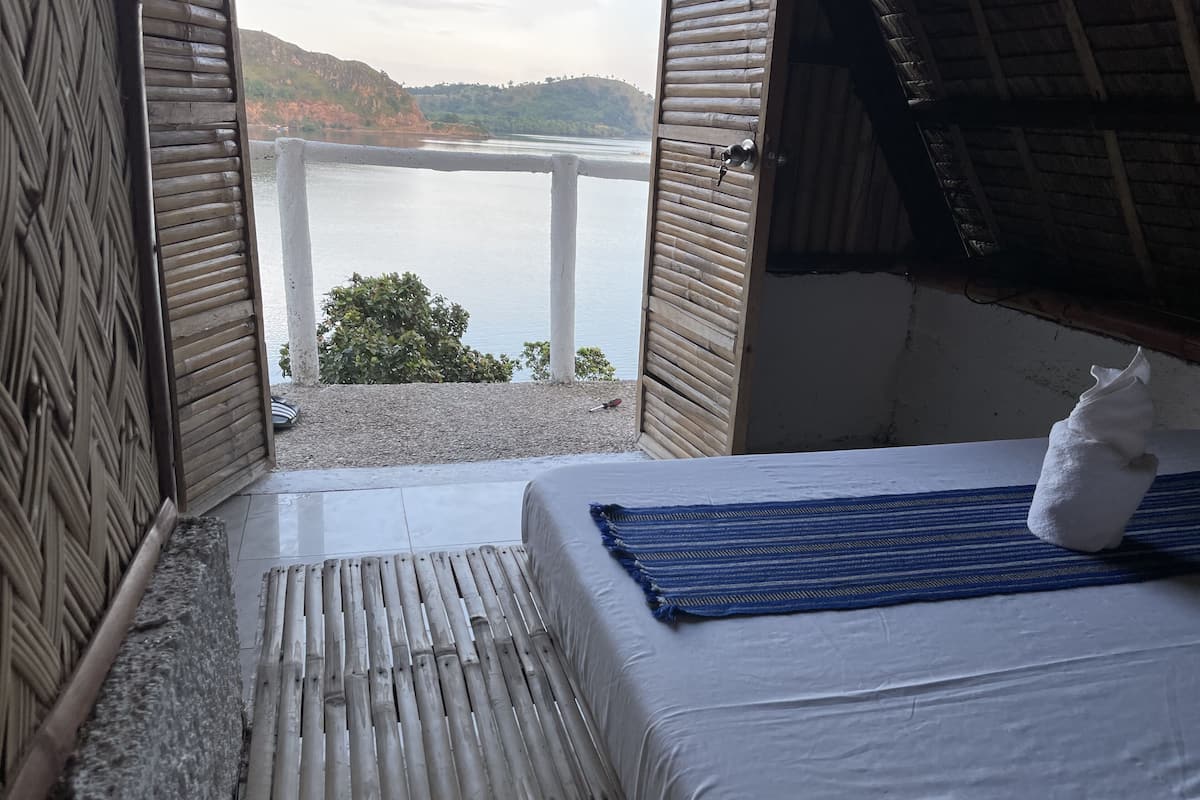 Una habitación con una cama, vista a un lago y techo de paja.