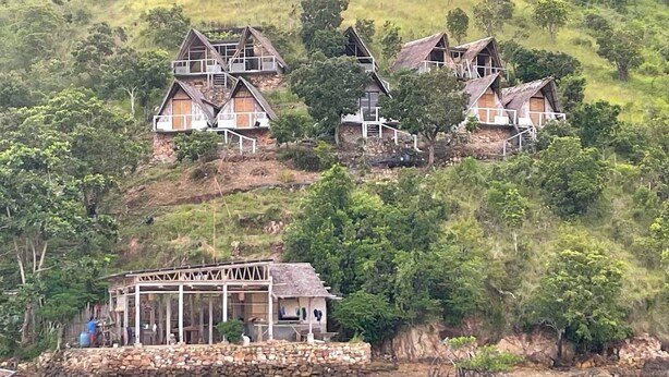 Eine Gruppe traditioneller Häuser an einem Hang, mit einer Steinmauer und einem hölzernen Steg, der sich ins Wasser erstreckt.