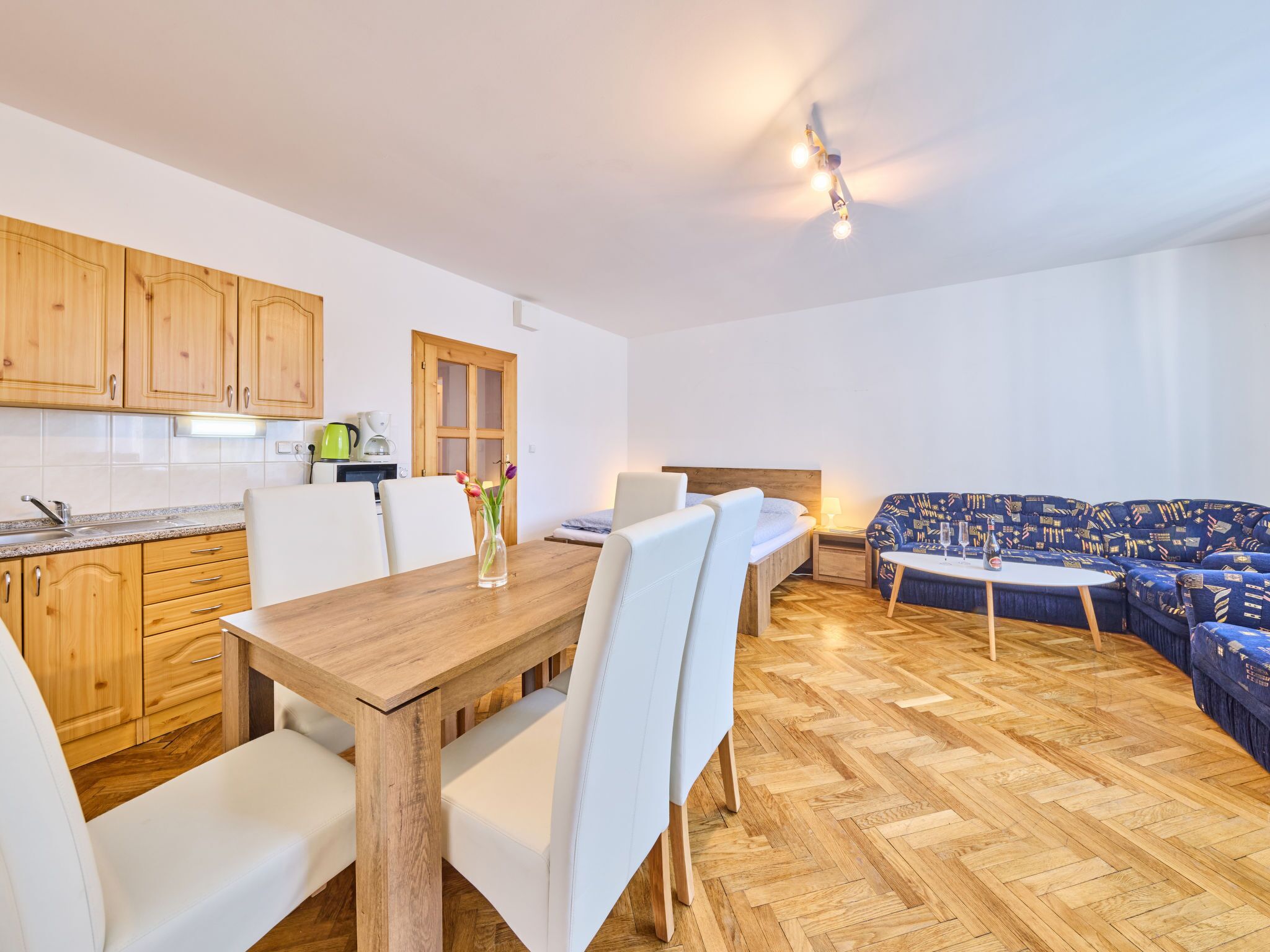 Una cocina moderna con armarios de madera y una mesa de comedor con sillas blancas. Hay un sofá y una mesita con una lámpara.