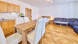 A modern kitchen with wooden cabinets and a dining table with white chairs. There is a sofa and a small table with a lamp. - Villa Belvedere by Interhome (Janské Lázně)