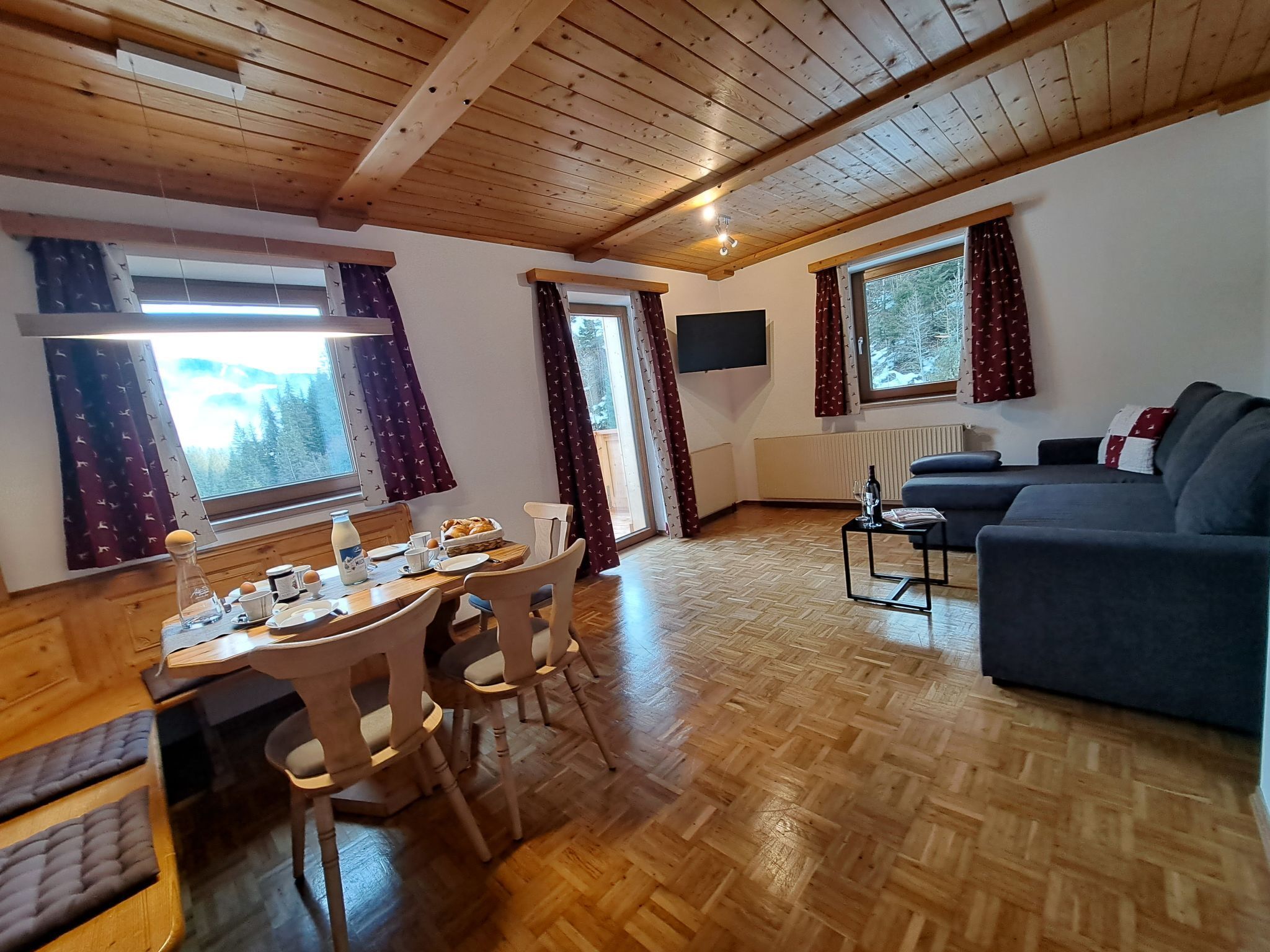 Een gezellige woonkamer met een houten plafond, een bank, een eettafel met stoelen en grote ramen met gordijnen.